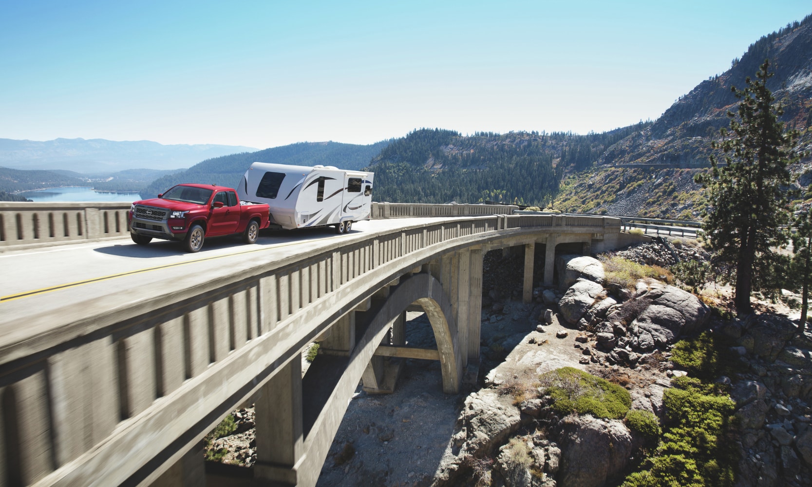 Image of a red 2025 Nissan Frontier towing a travel trailer over a bridge in a mountainous lake landscape during the day