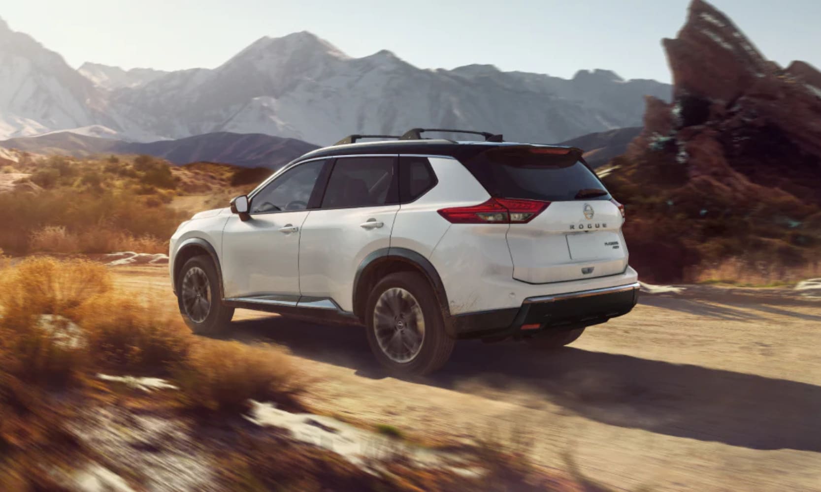 Exterior rear side image of a white 2025 Nissan Rogue driving on a dirt road in a mountainous landscape
