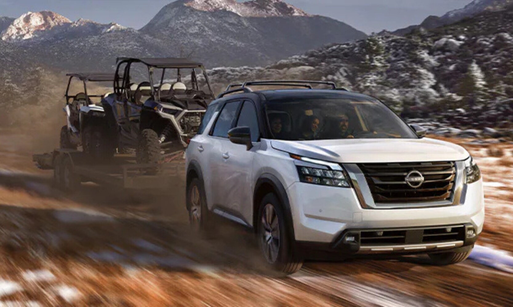 Exterior front image of a white 2025 Nissan Pathfinder towing two ATVs in a rocky mountain landscape
