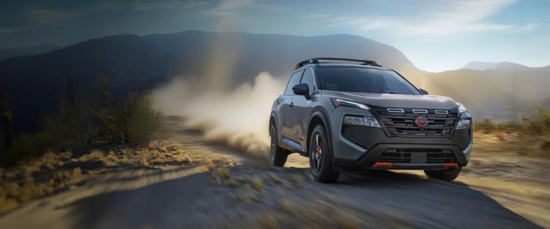 Hero image of a tan 2025 Nissan Rogue driving on a dirt road with dirt blowing behind the tires and mountains in the background