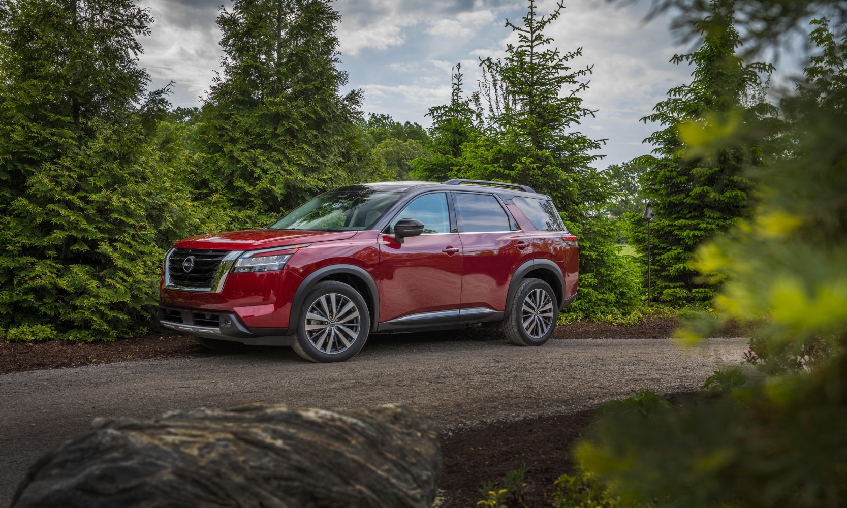 Exterior side image of a red 2024 Nissan Pathfinder Platinum parked on the side of a tree-lined dirt road
