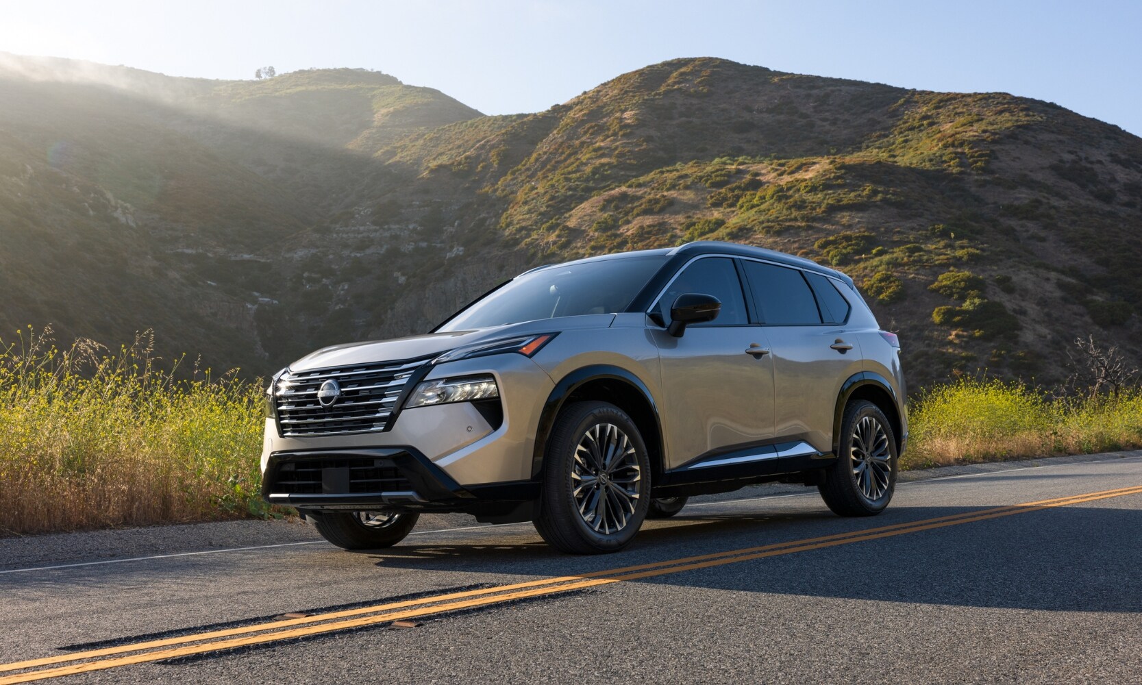 Exterior side angle image of an off-white 2024 Nissan Rogue driving through a mountainous road during the day