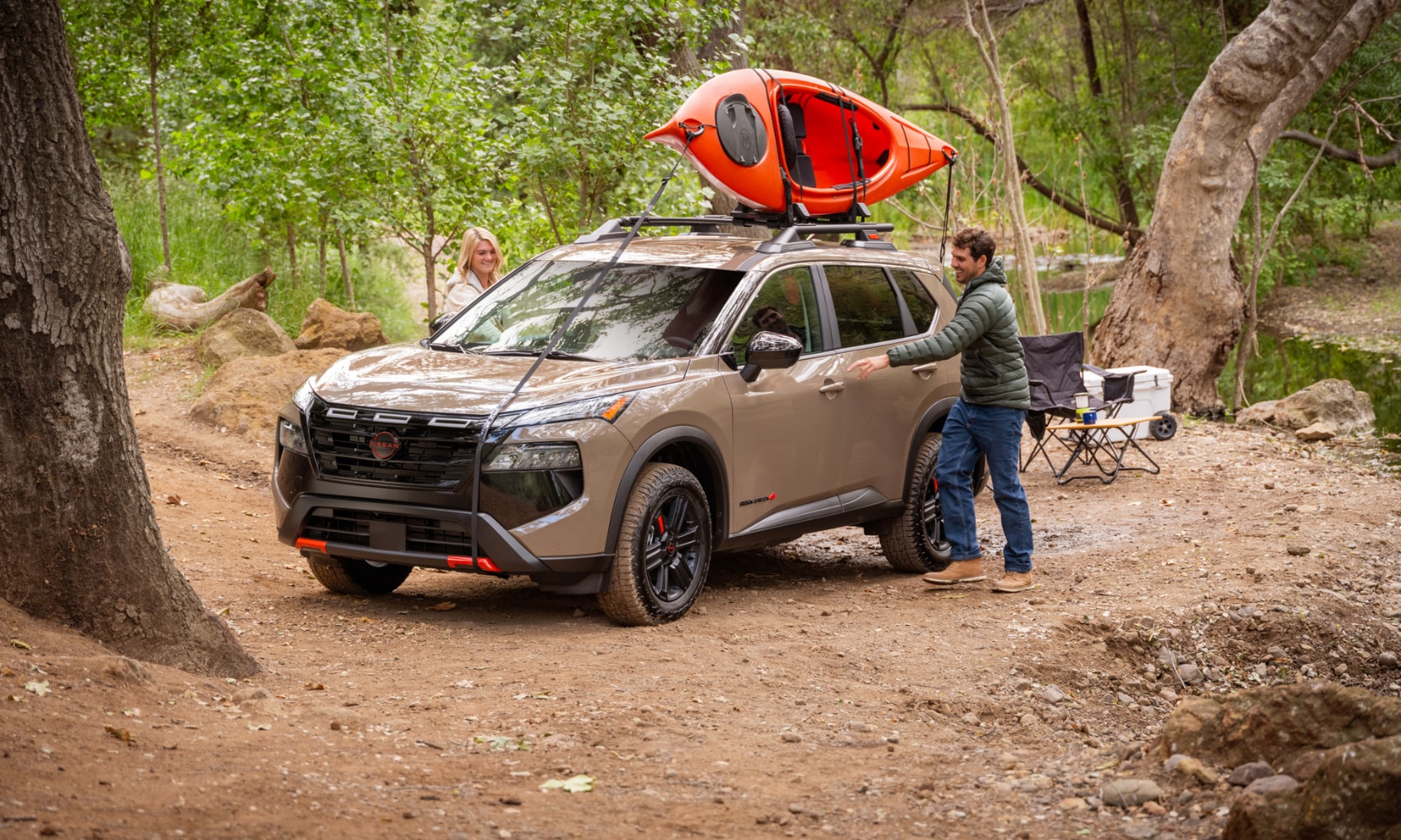 Exterior image of two people entering a tan 2025 Nissan Rogue Rock Creek in a forest with a kayak tied to the roof