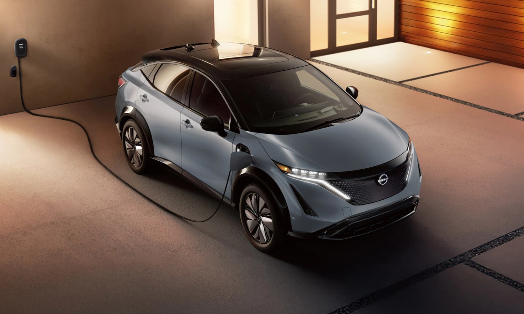 Top angle exterior image of a light blue 2025 Nissan ARIYA charging at a home's driveway at night
