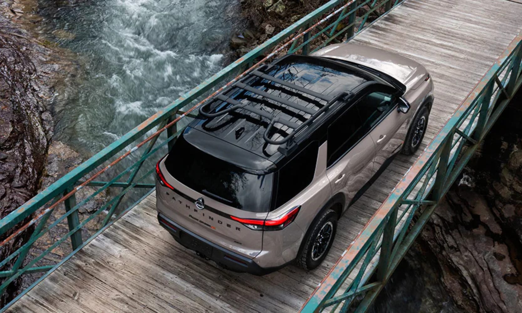 Top-view exterior image of a taupe 2025 Nissan Pathfinder Rock Creek parked on a bridge over rocky water cliffs