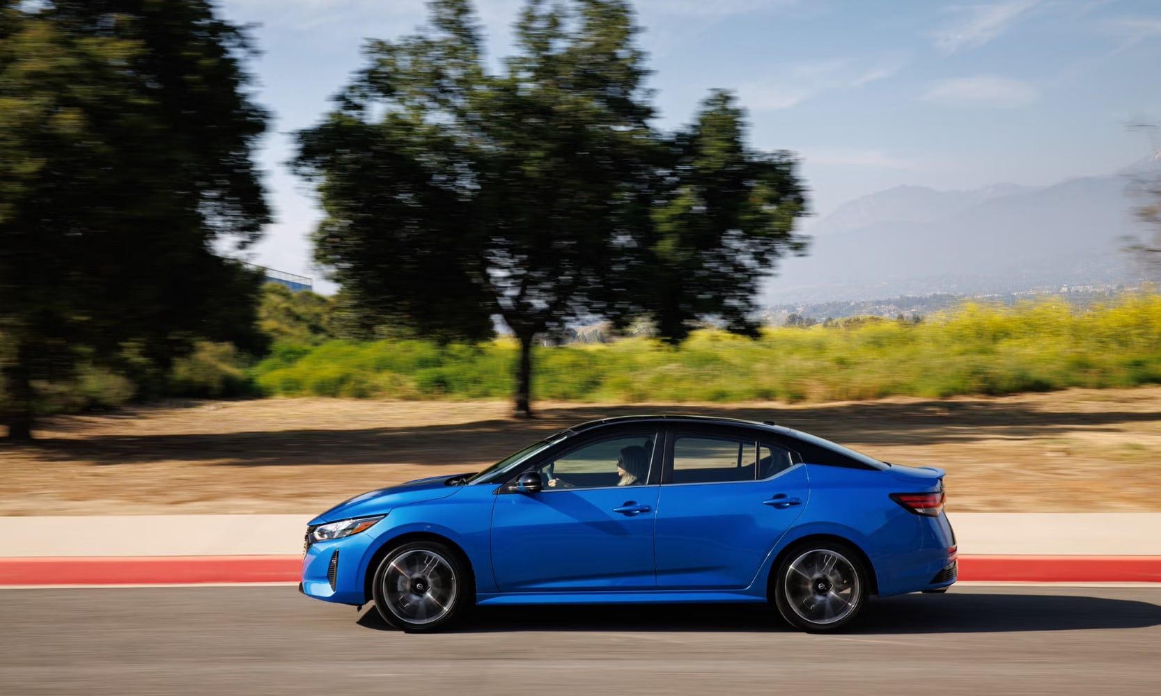 Exterior side image of a blue 2024 Nissan Sentra SR driving down a tree-lined street with mountains in the background