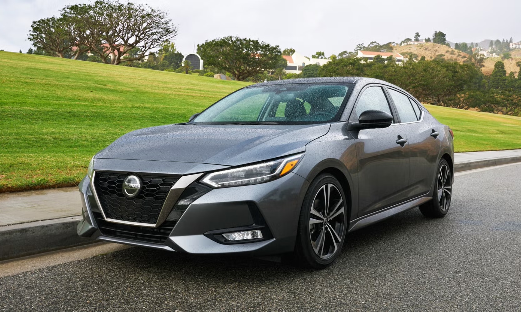 Exterior front angle image of a gray 2020 Nissan Sentra parked on a suburban street with mountains in the background