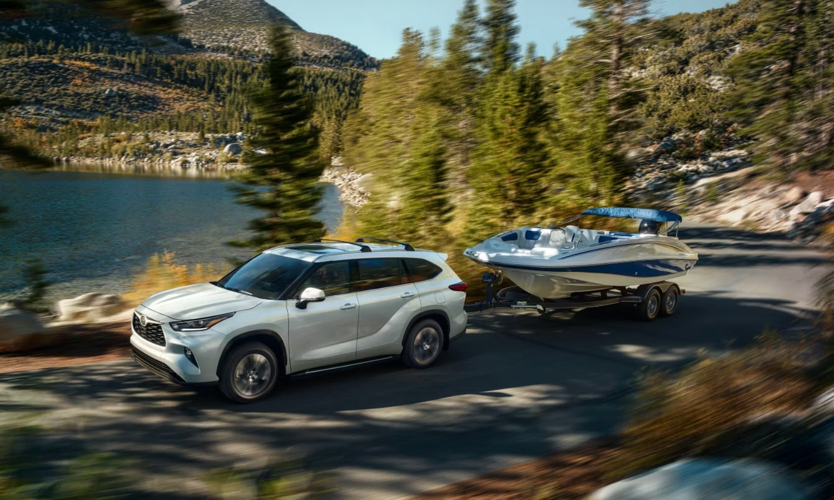 Image of a white 2025 Toyota Highlander towing a small bout through a mountainous road with a lake in the background