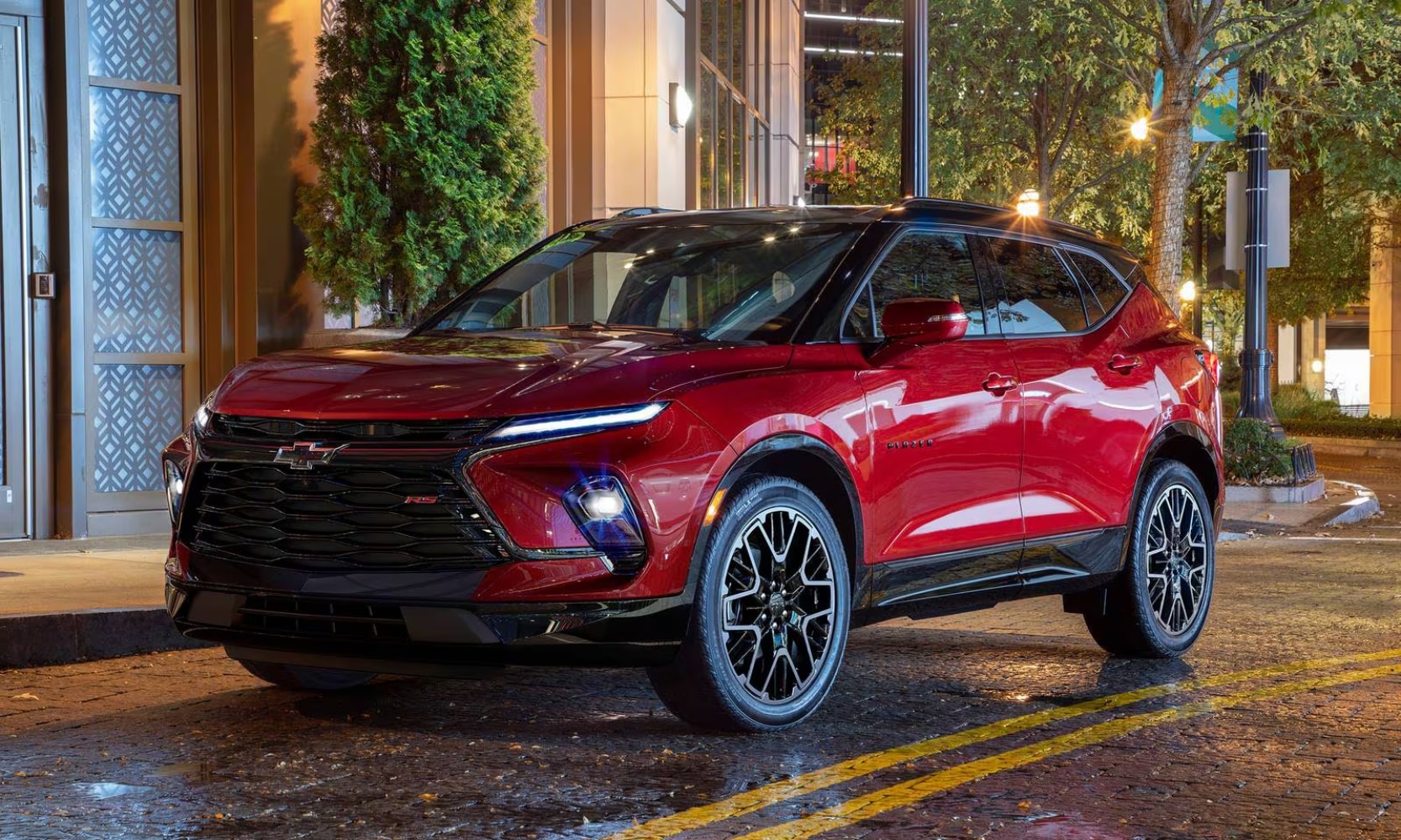 Exterior image of a red 2023 Chevy Blazer parked on a city street in the rain with the lights on