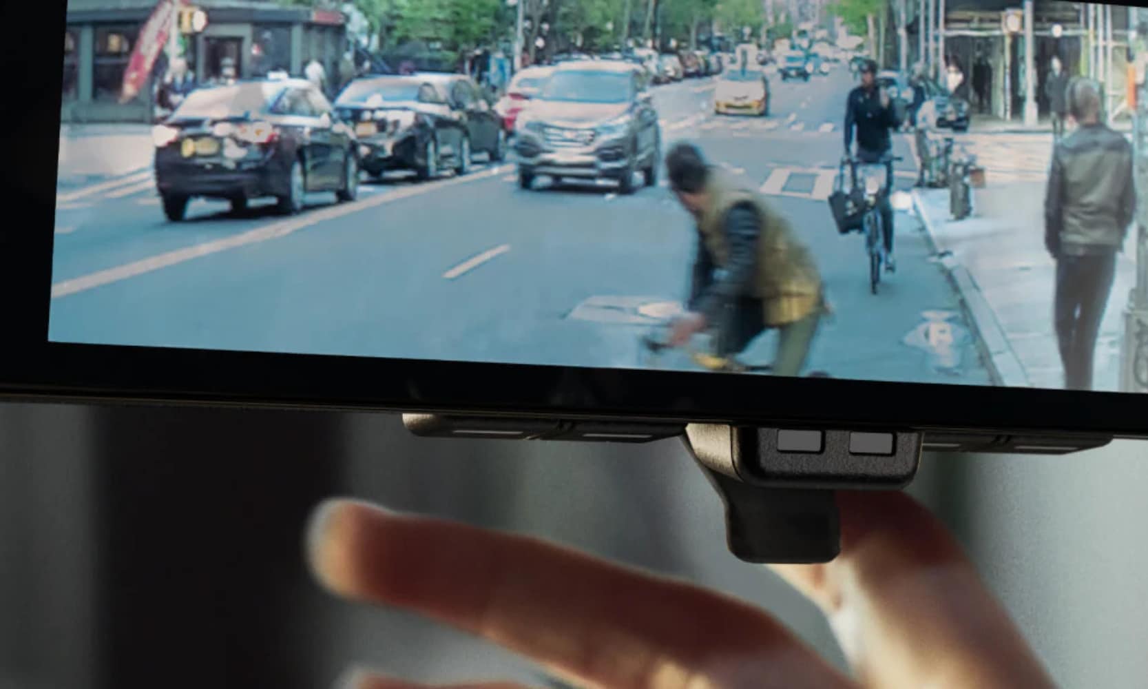Closeup image of a hand touching the 2025 Nissan ARIYA Rear View Camera showing vehicles and pedestrians on the street