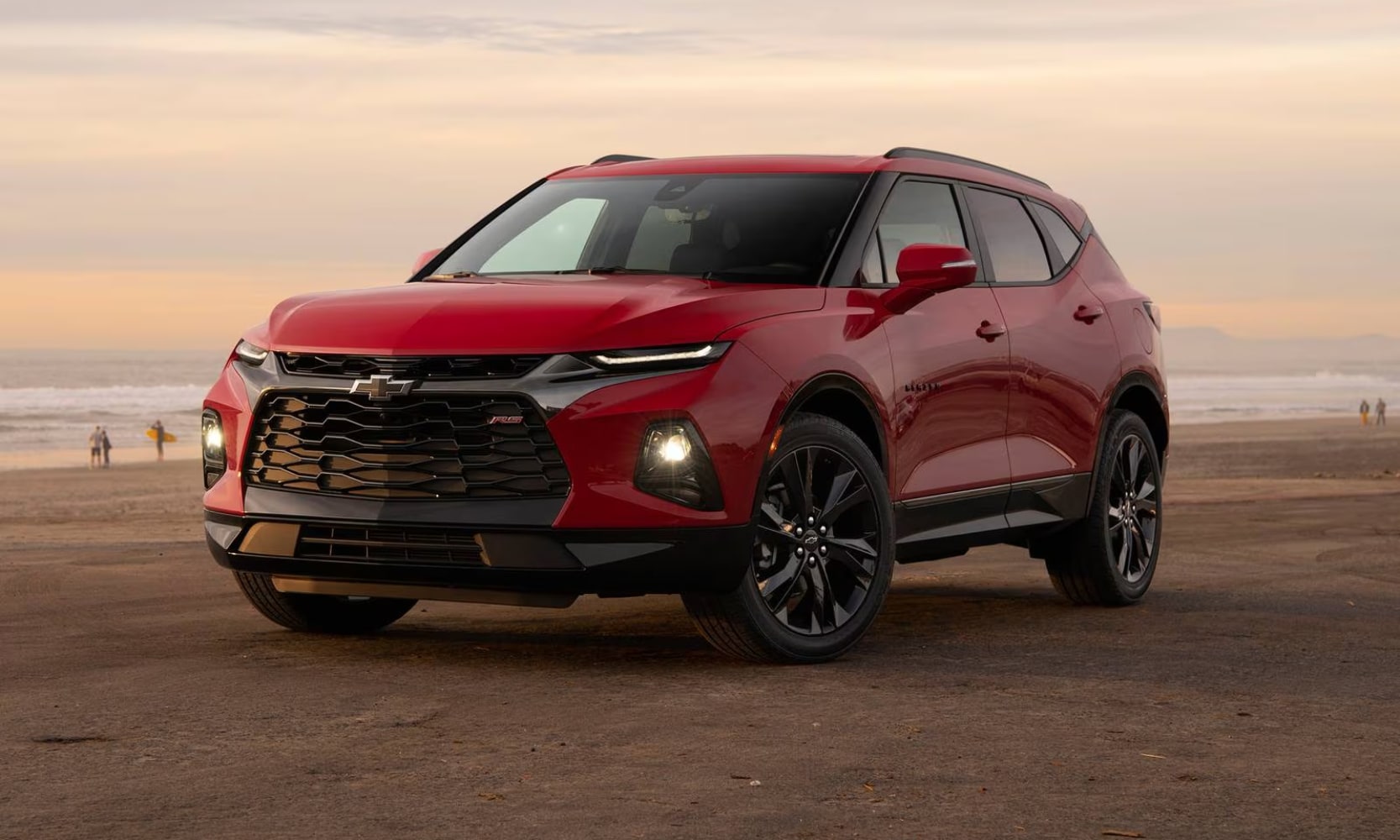 Exterior image of a red 2021 Chevy Blazer parked on a beach with its lights on during sunset with people in the background