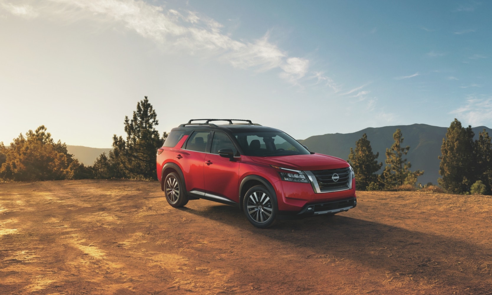 Exterior image of a red 2025 Nissan Pathfinder Platinum parked on a hill with forest trees and mountains in the background