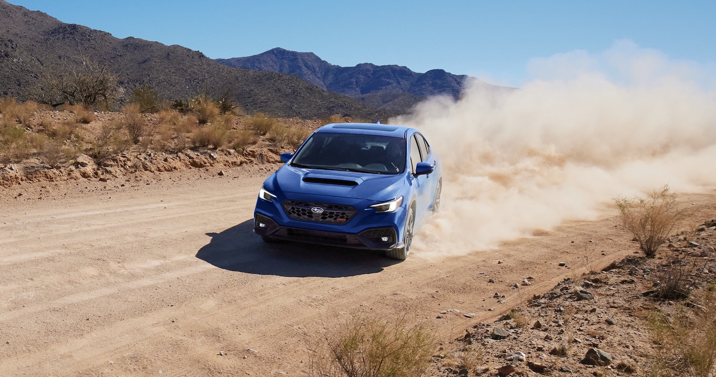 A blue 2025 WRX 4-door sports car kicks up a large cloud of dust rounding a corner on a gravel road.