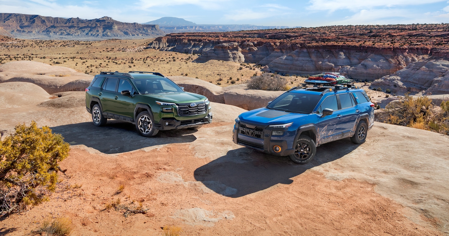  A 2026 Subaru Outback in emerald green and a 2026 Subaru Outback Wilderness in geyser blue parked next to each other on a mountain overlooking a desert.