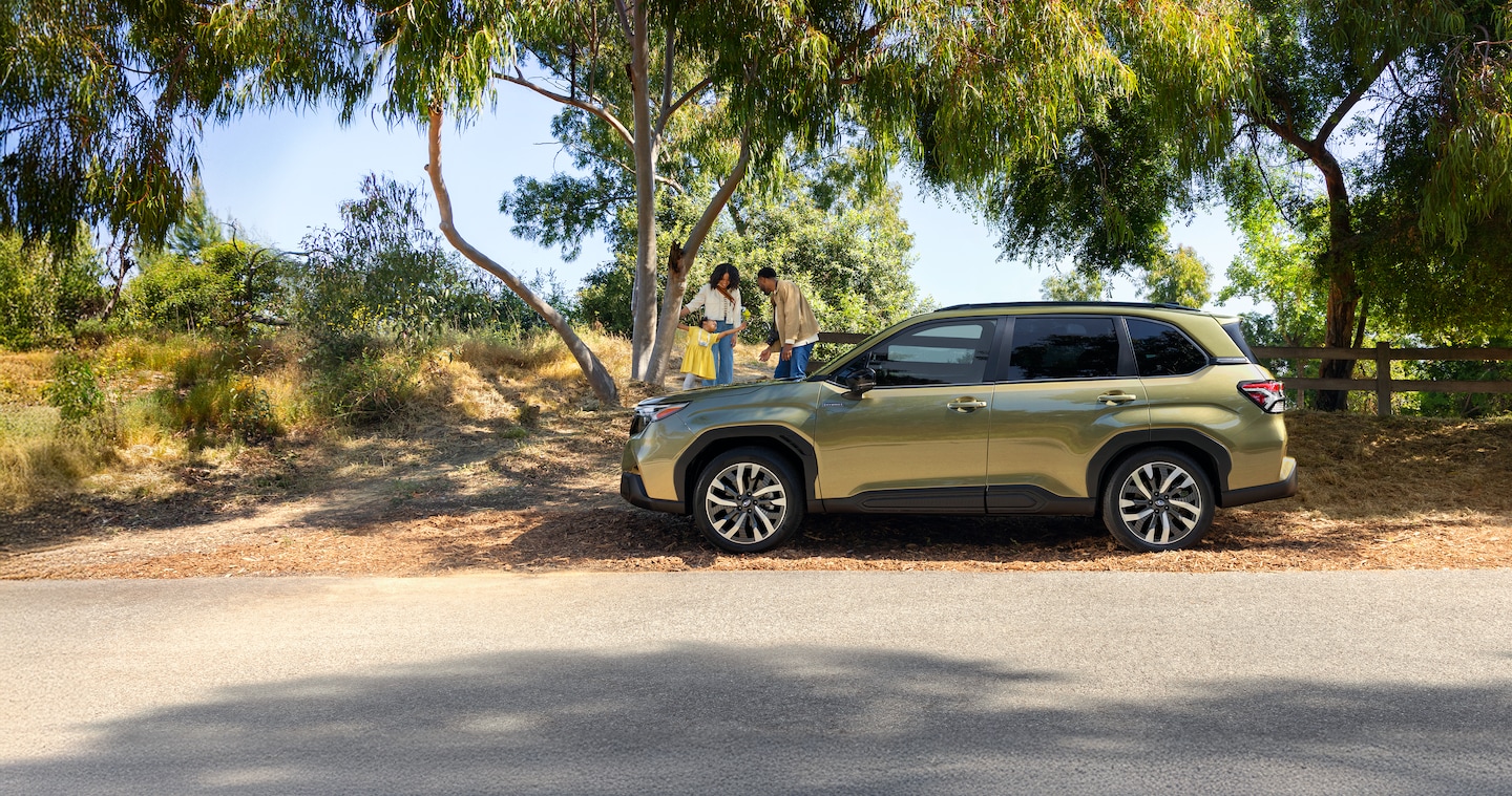  A 2026 Forester Hybrid in Autumn Green Metallic parked on the road as a family plays with their daughter in the background.