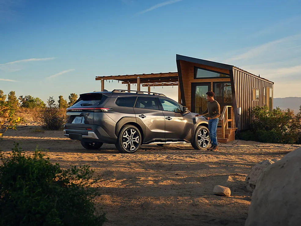 A man charges his Subaru Trailseeker next to a small home.