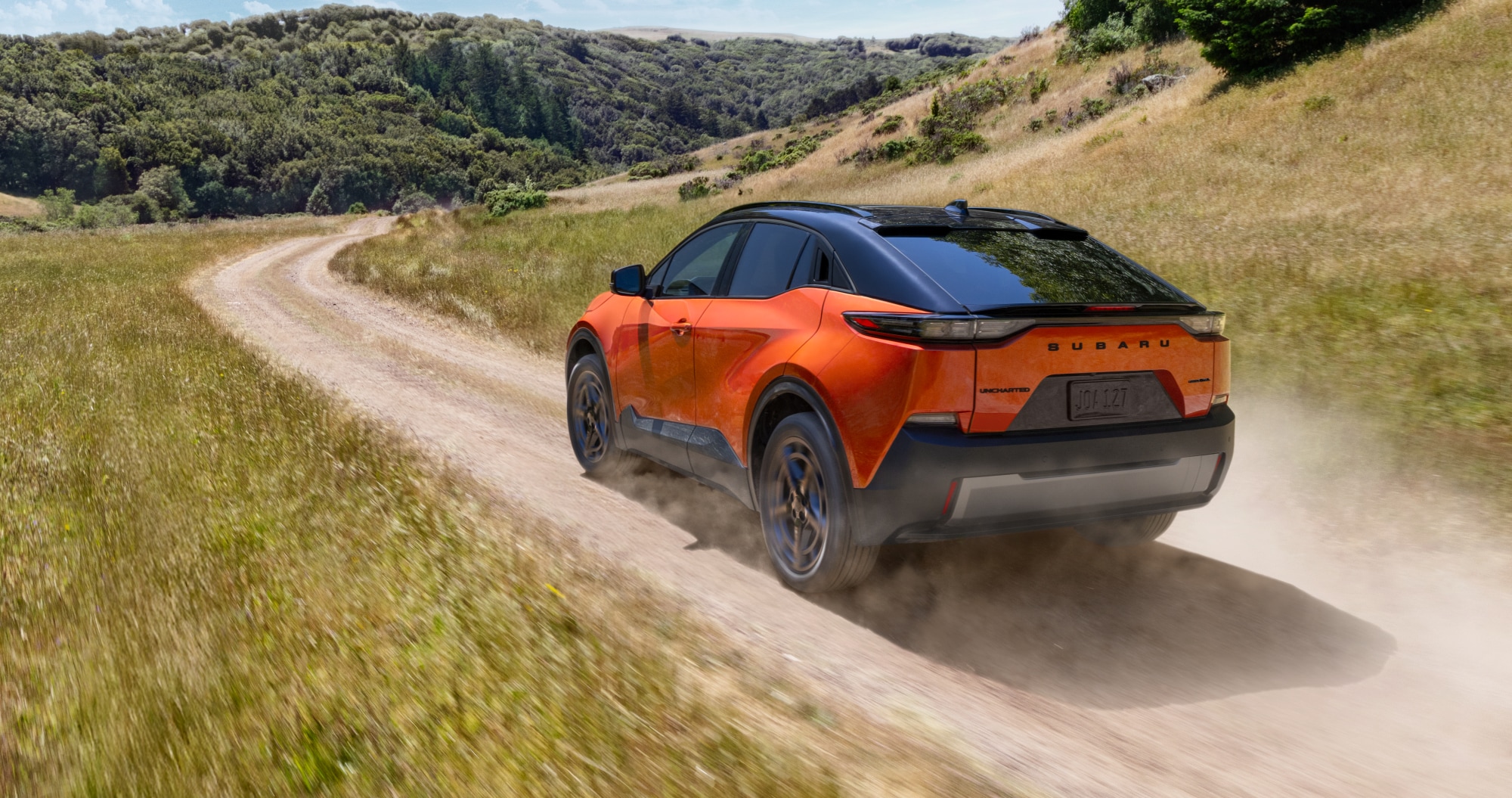  A static image of an orange 2026 Subaru Uncharted driving on a winding dirt road through wide, grassy hills. 