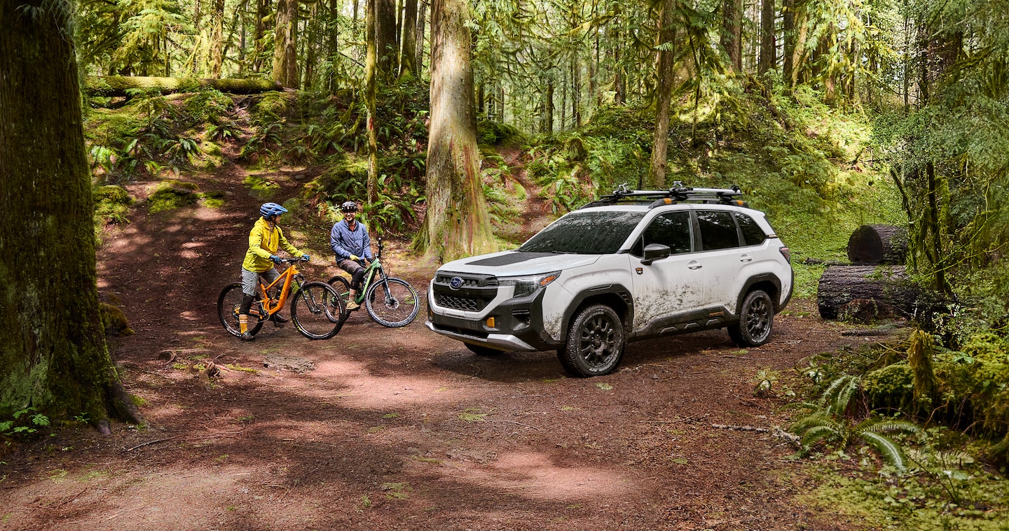 Two people on off-road bicycles next to a muddy 2026 Subaru Forester Wilderness in white in the middle of a forest.