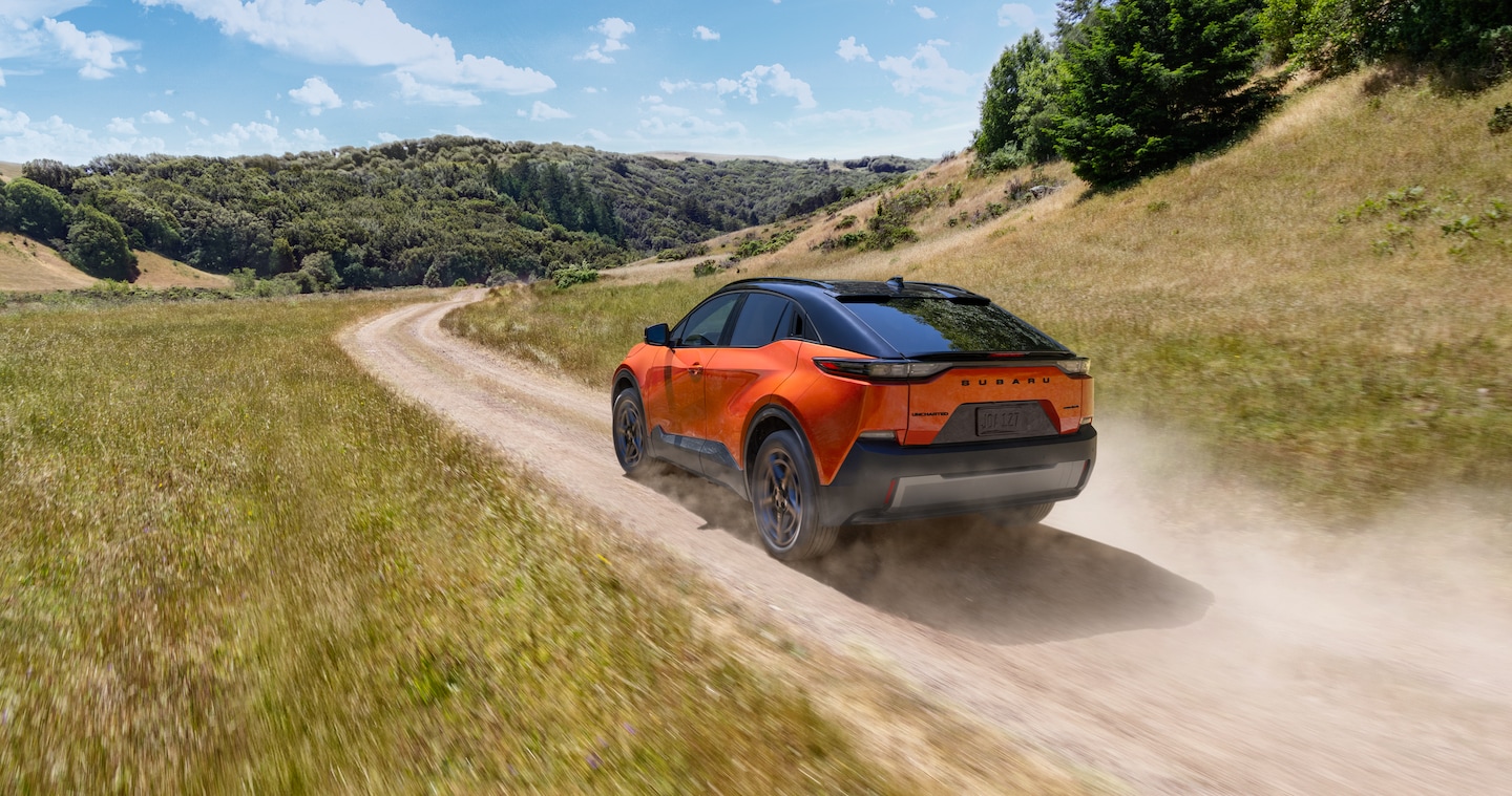  An orange Subaru Uncharted electric SUV drives on a dirt road through some rolling hills.
