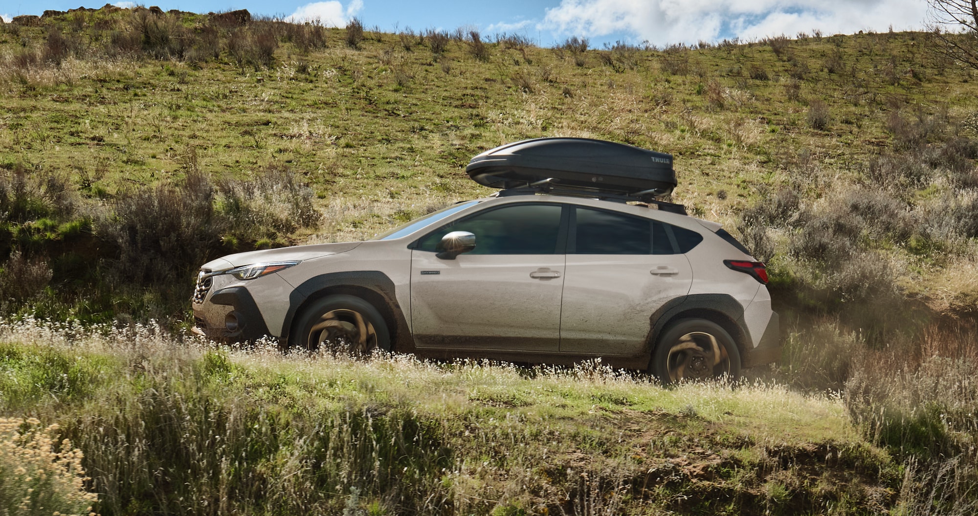  A beige Subaru Crosstrek Hybrid drives off‑road through a grassy, uneven hillside with a rooftop cargo box attached under a bright, partly cloudy sky.