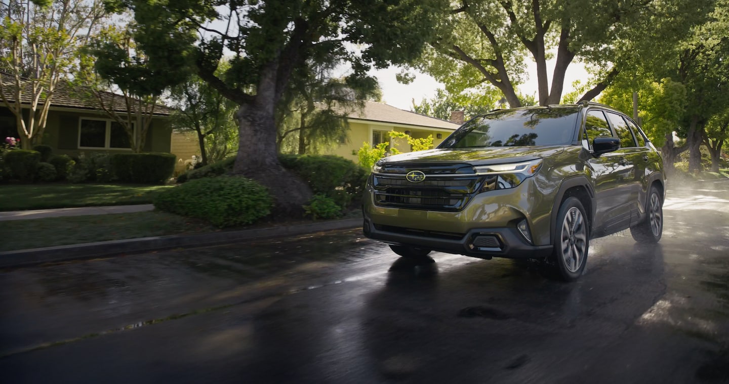 2025 Subaru Forester Hybrid driving on a wet, tree-lined neighborhood street.
