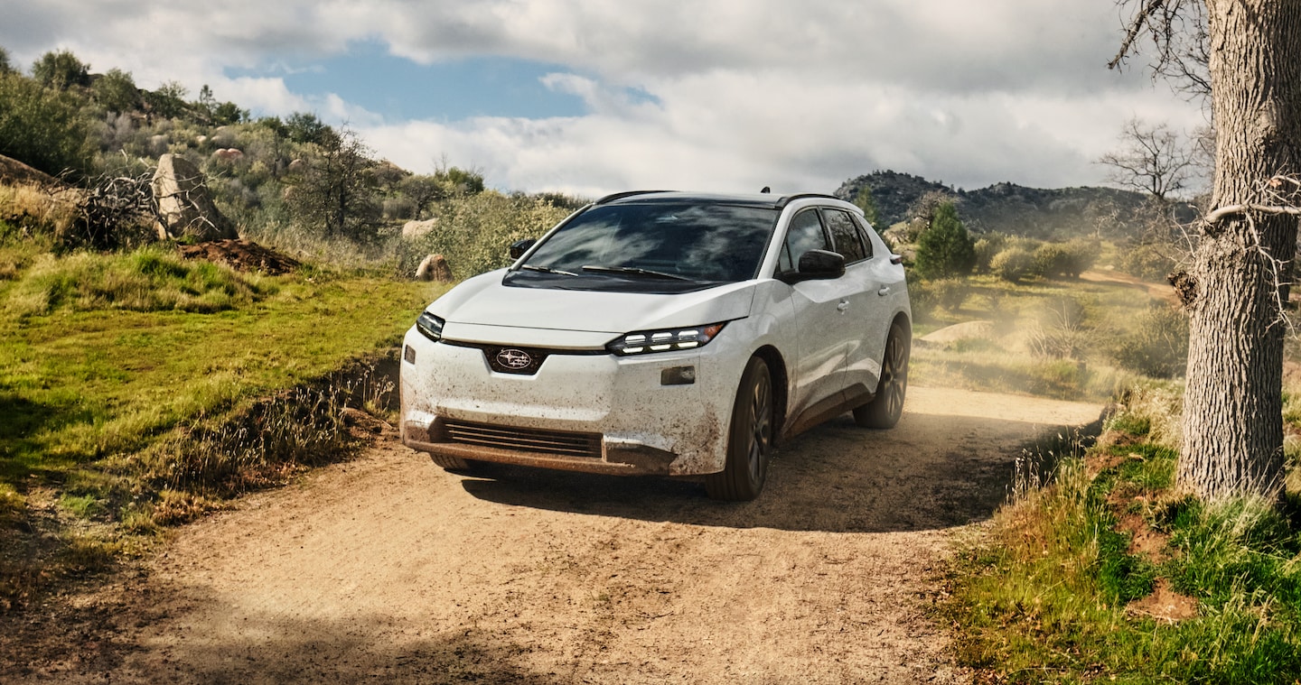  A white 2026 Subaru Solterra electric SUV drives on a dirt road in arid hills, kicking up dust behind.