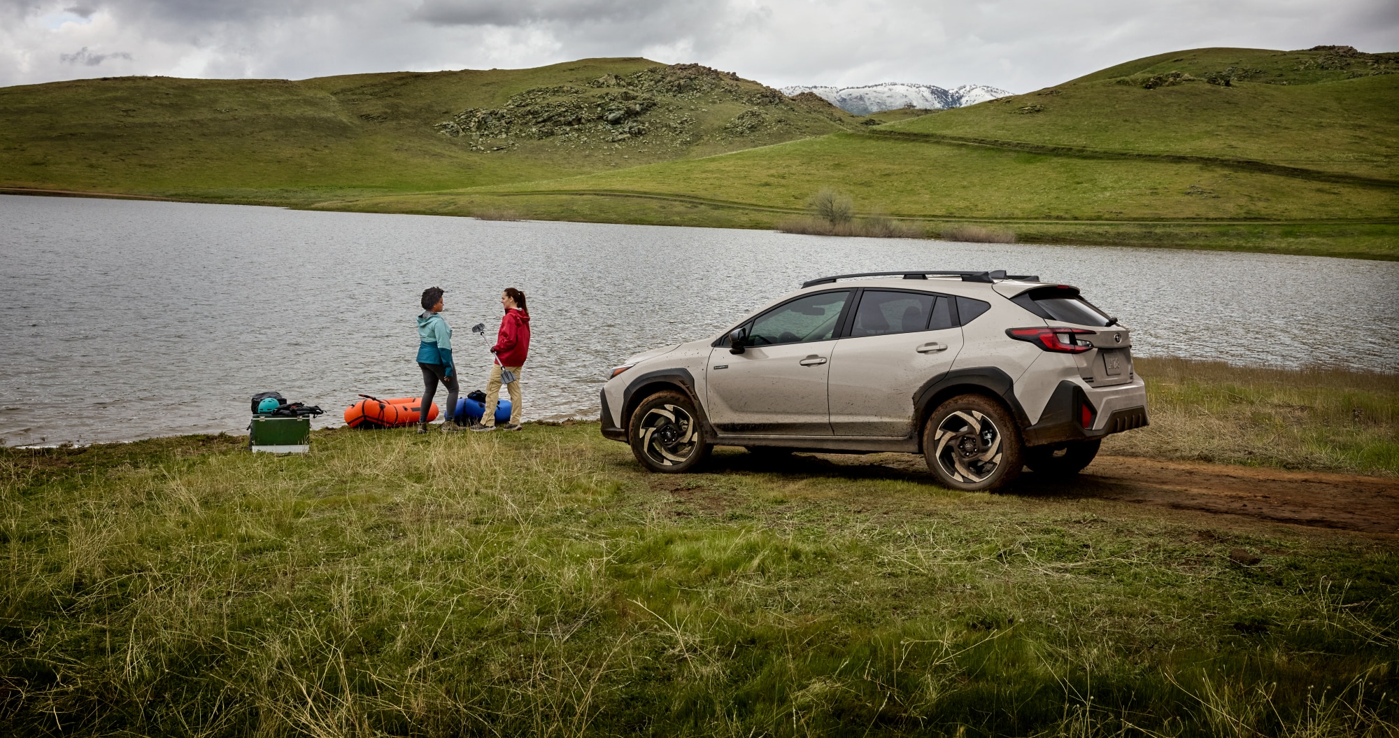  A Subaru Crosstrek Hybrid parked beside a lakeshore on a grassy field, with two people standing near camping gear by the water under a cloudy sky and rolling green hills.[