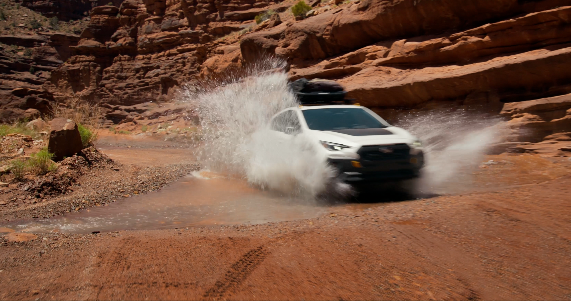 A 2027 Crosstrek Wilderness off-road compact SUV splashes through a puddle in a canyon.  
