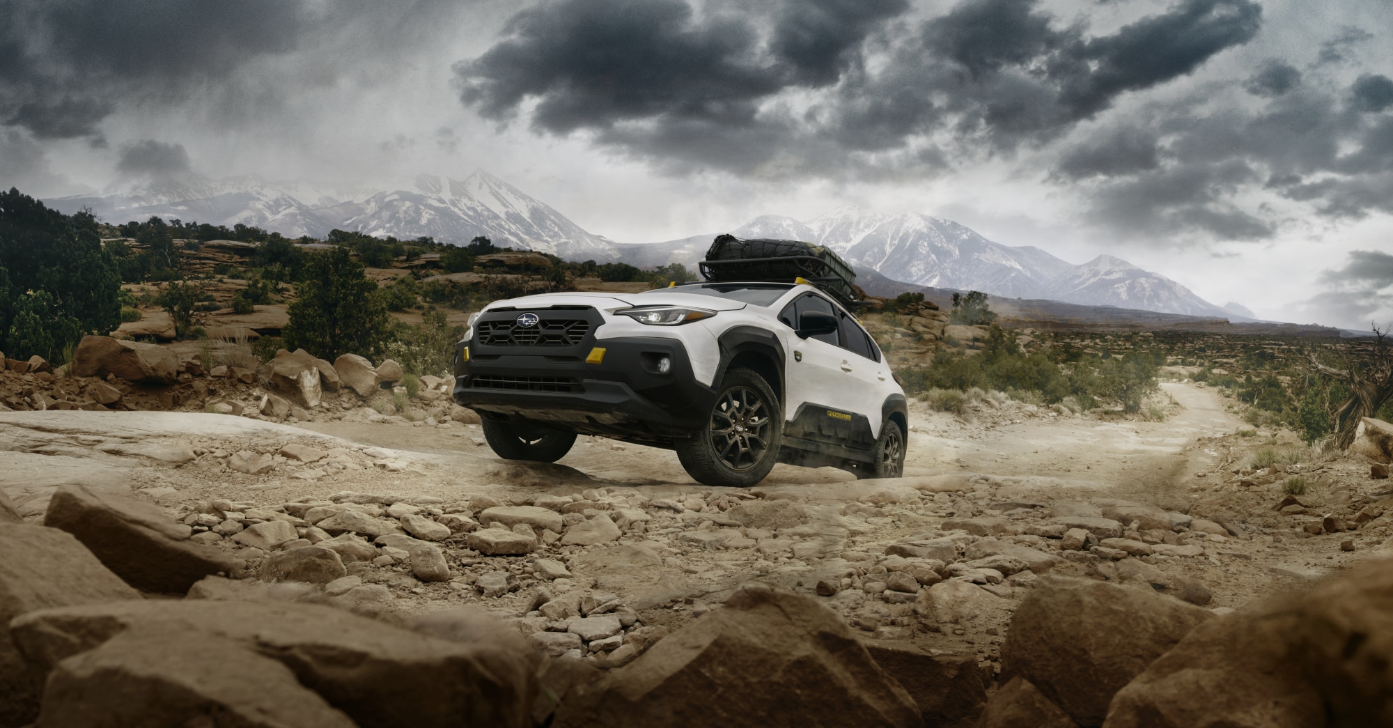  A static image of a 2026 Crosstrek Wilderness driving over a rocky dirt trail in a rugged, mountainous landscape under dark, stormy clouds. 