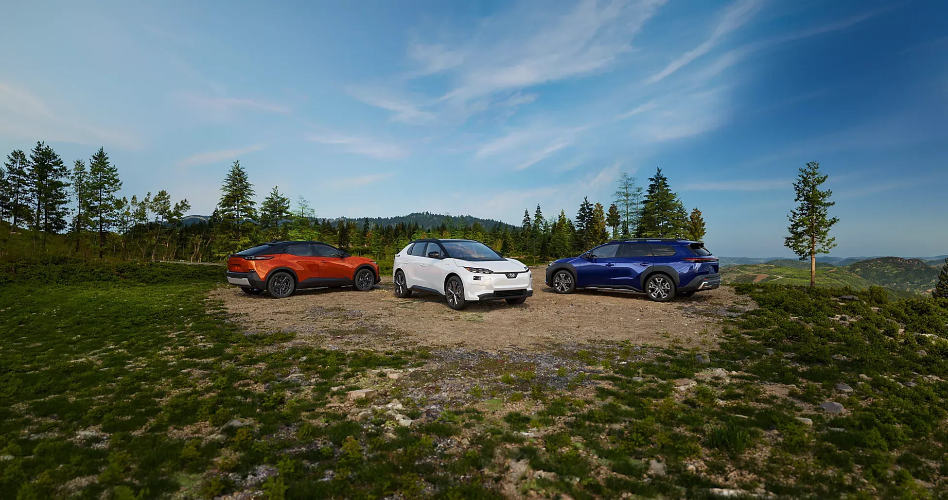 Three Subaru SUVs, one orange, one white, and one blue, are parked on a gravel clearing surrounded by evergreen trees. The vehicles are positioned side by side against a backdrop of rolling forested hills beneath a bright blue sky with wispy clouds. The foreground features rocky, grassy terrain, emphasizing an outdoor and scenic setting.