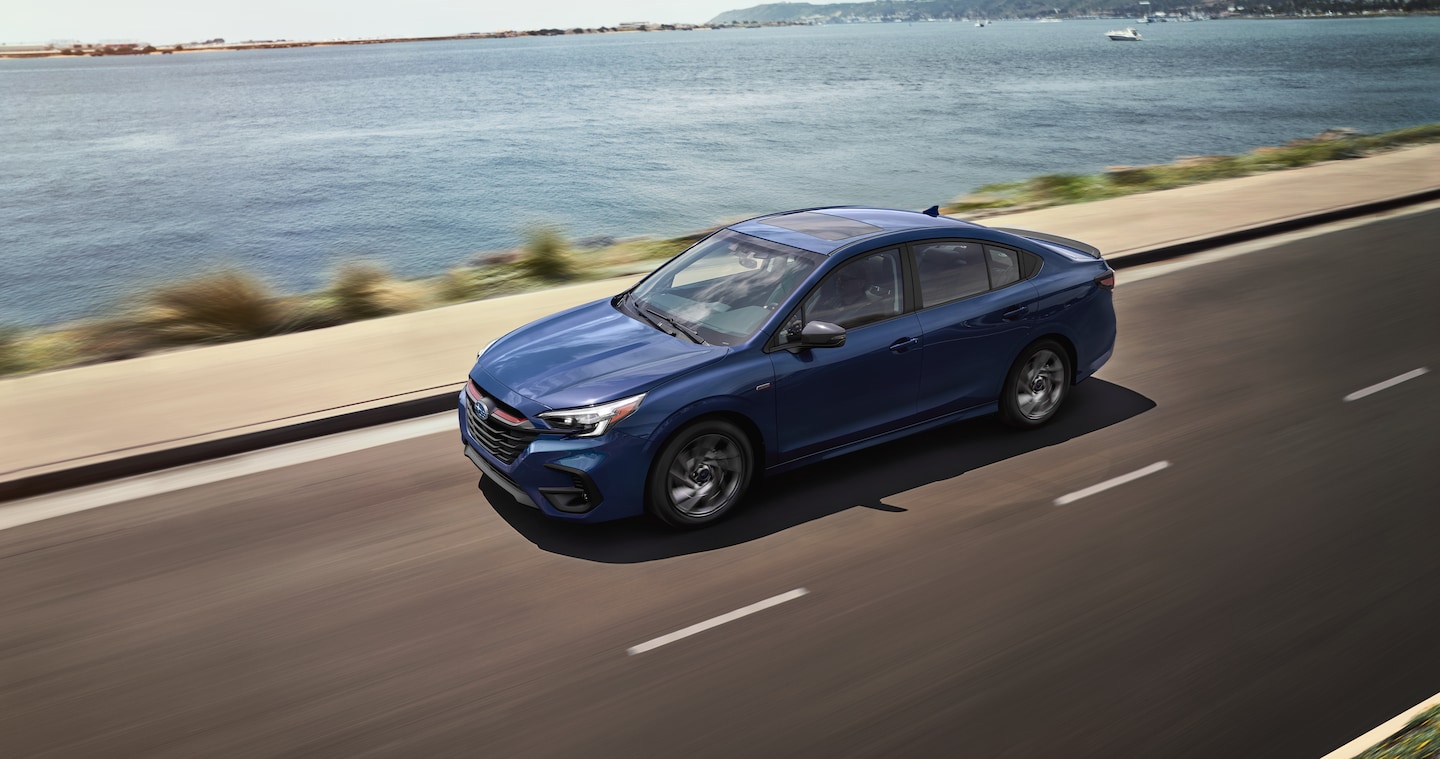 A blue Subaru Legacy sedan drives next to the ocean.