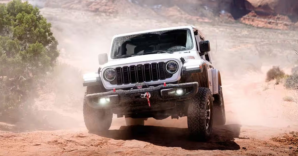A white 2026 Jeep Wrangler Rubicon kicking up dust while navigating a rugged, dirt trail in a rocky desert.