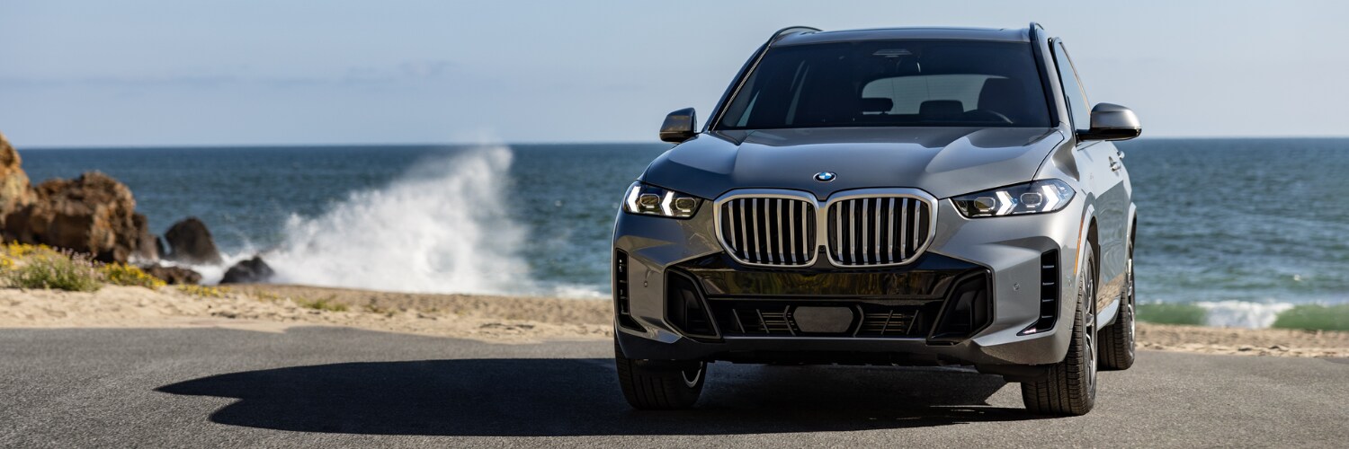BMW X5 PHEV silver BMW X5 PHEV SUV parked on the beach with a wave crashing in the background