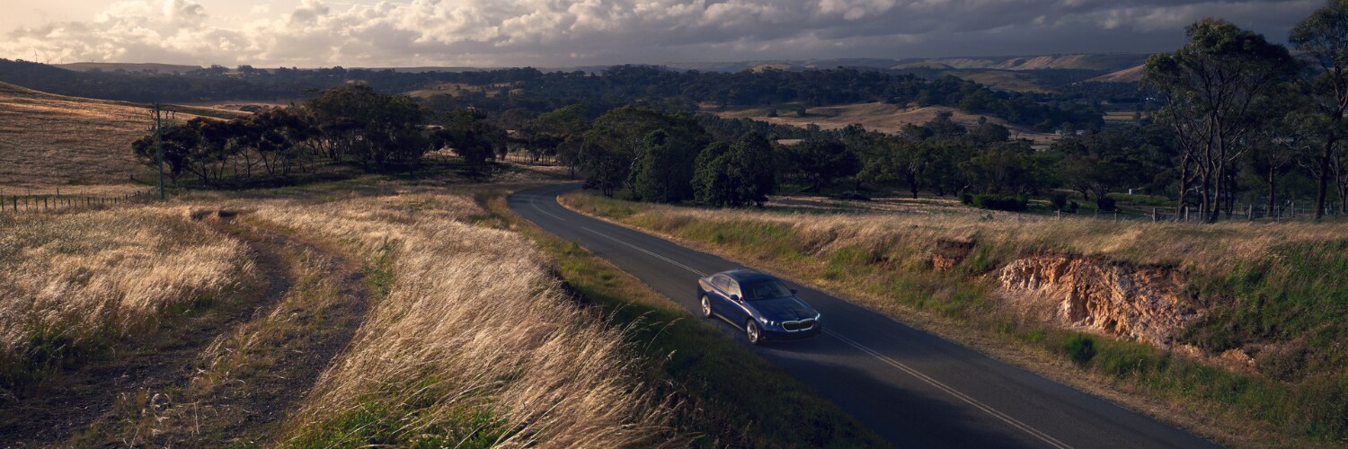 dark blue BMW 5 Series sedan driving on a road through a grassy field