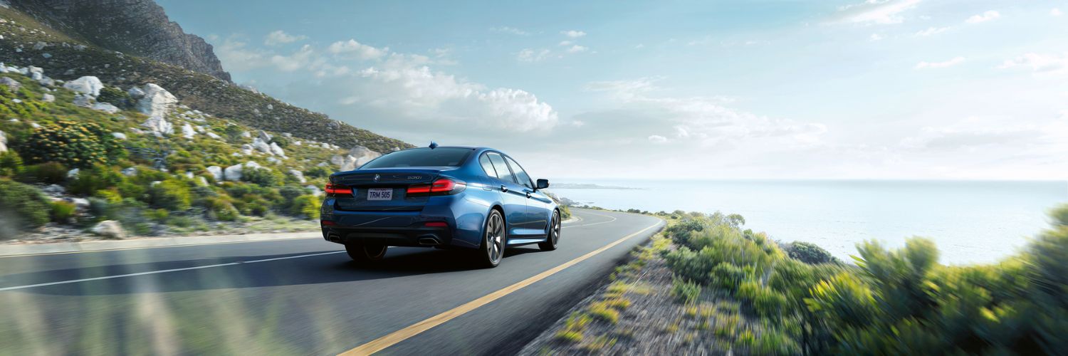 blue BMW m550i 5 Series sedan driving on a curving road near the water