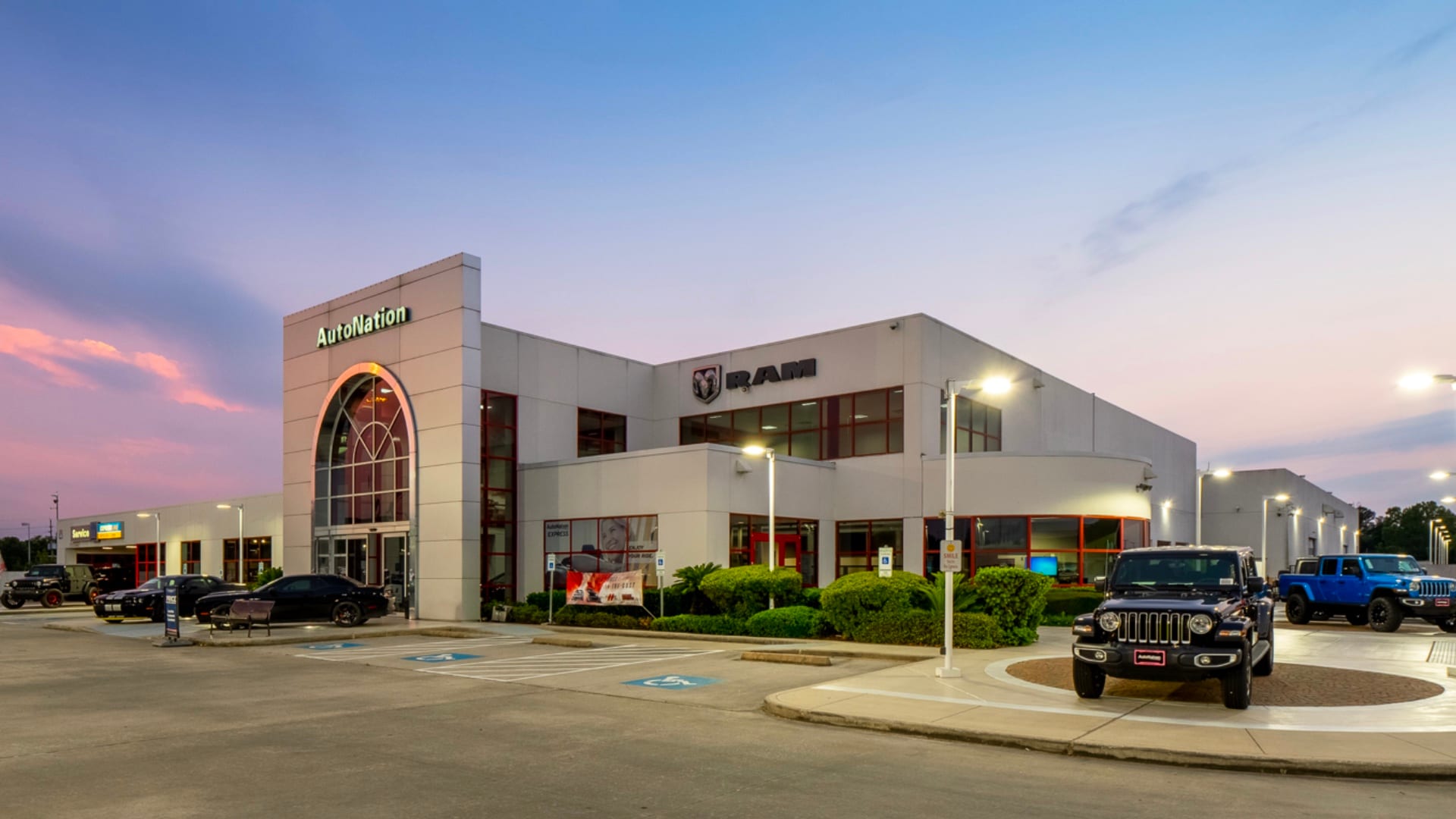 Exterior view of AutoNation Chrysler Dodge Jeep Ram Spring during a sunrise or sunset. Many vehicles parked near the grey building. Trees and greenery are visible around the parking lot and surrounding area.