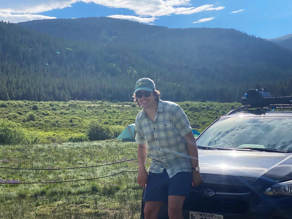 Jack, wearing a Stanley Subaru hat, on his Crosstrek in front a beautiful view of a lea and low mountain