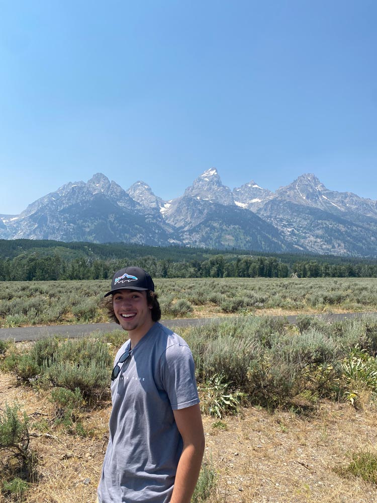 smiling guy in the foreground, meadow and woods in the midground, mountains in the background