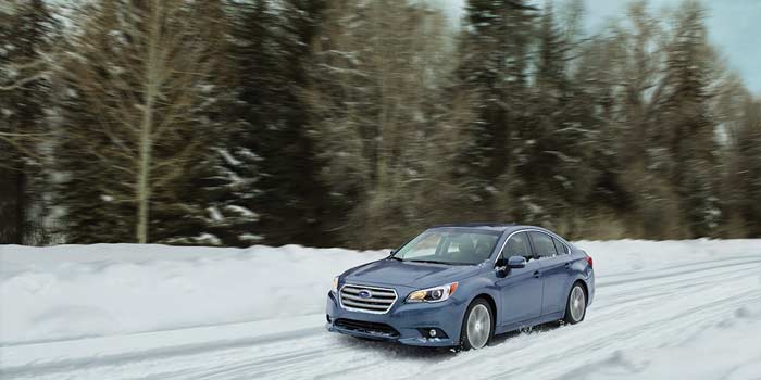 Subaru Legacy driving through the snow backed by tall conifers and blue sky