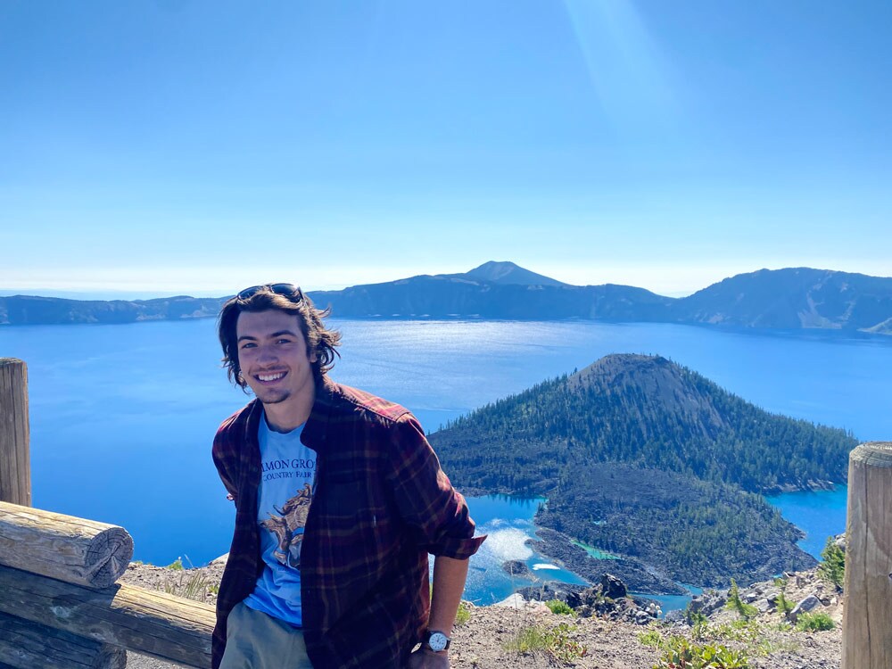 Smiling man leaning against a wood log rail with a large bay in the background featuring a large island
