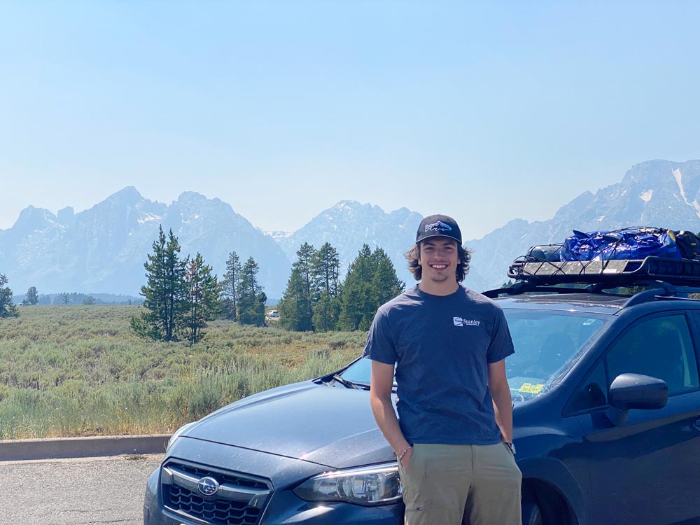 smiling guy leaning on his Crosstrek with mountains in the distant background, faded blue by atmospheric perspective