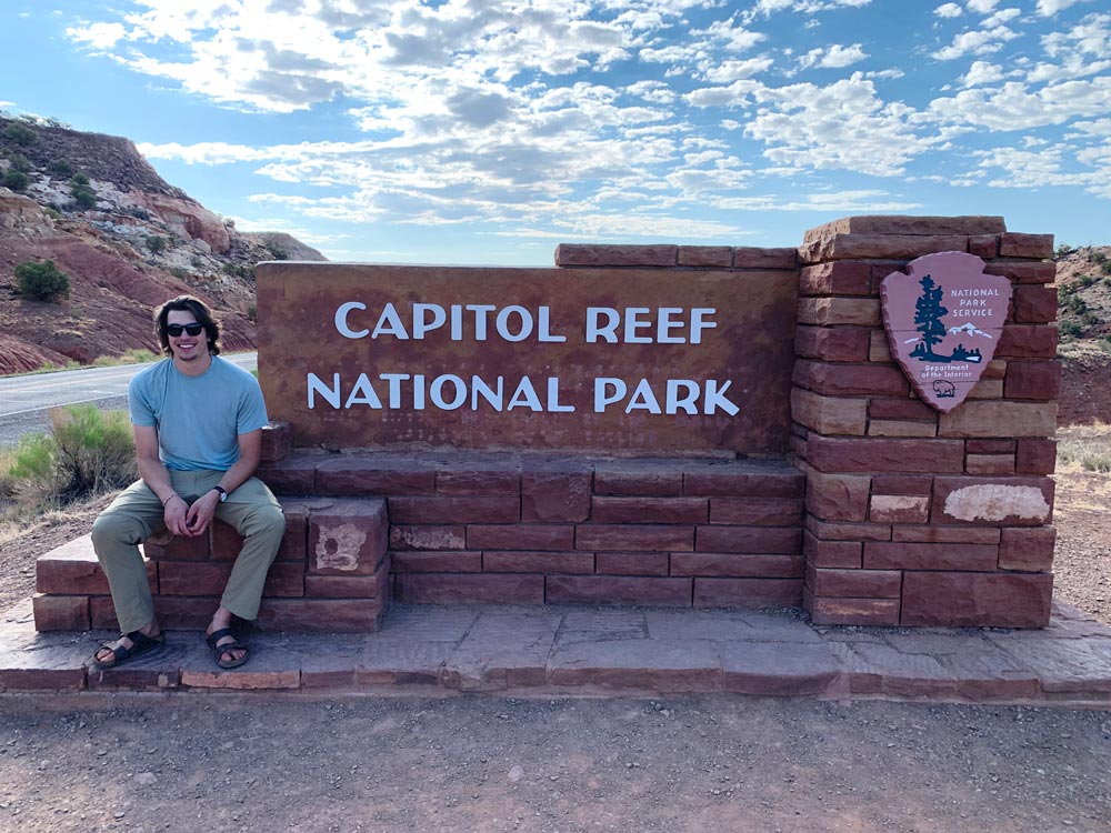 Smiling man sitting next to the large stone Capitol Reef National Park sign