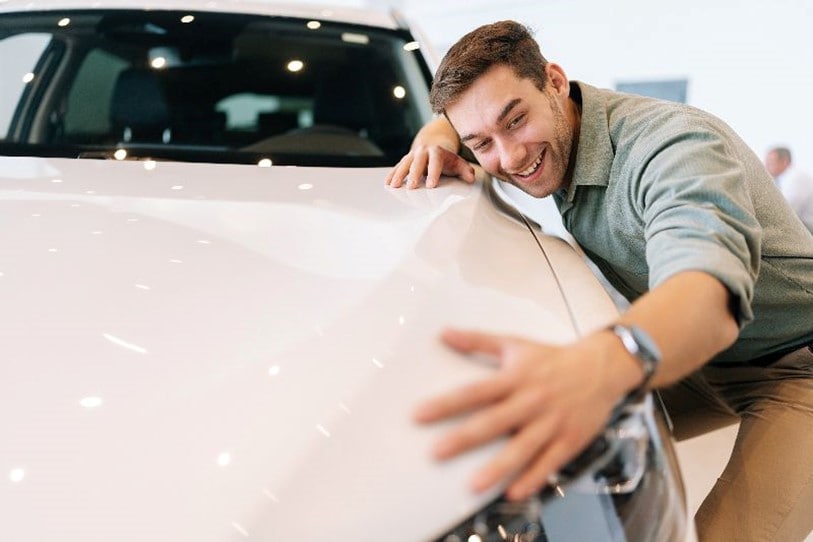 A man smiling and leaning against a car, touching its hood