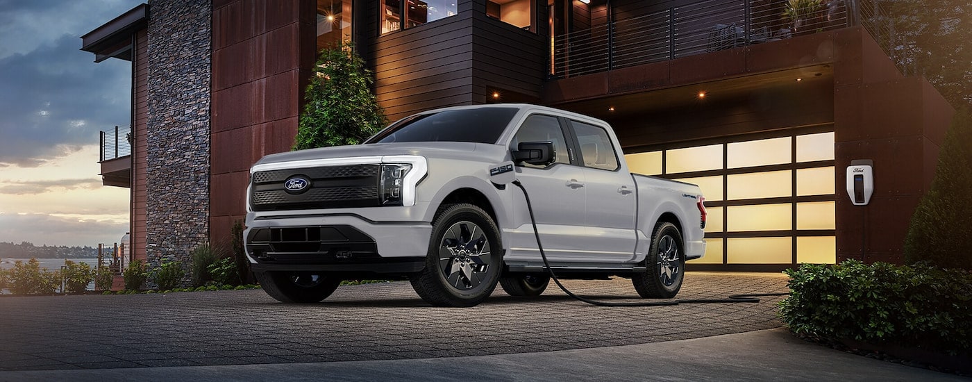 A white 2024 Ford F-150 Lightning charging in the driveway of a modern home.