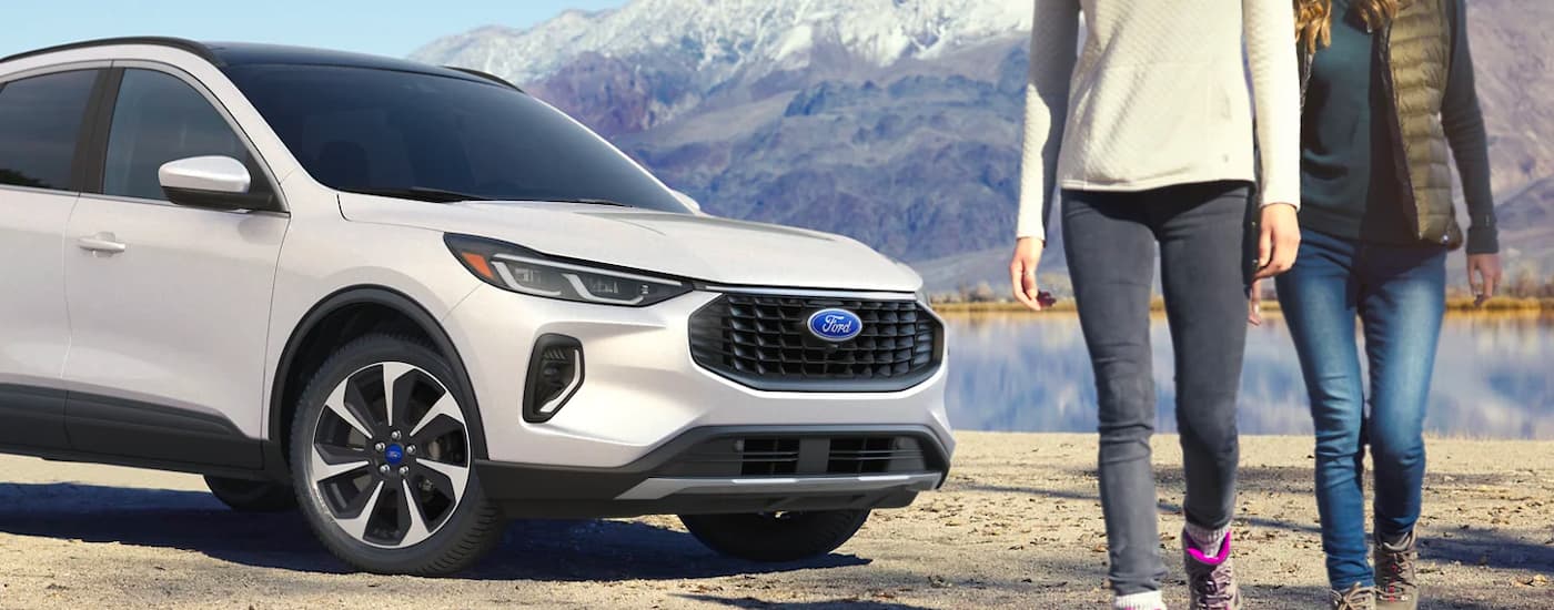 Two people walking from a white 2023 Ford Escape Platinum parked by a lake.