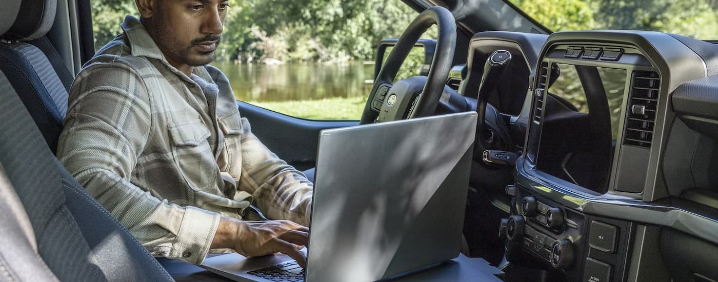 A man is using a laptop in the front seat of a 2025 Ford F-150 for sale near Trenton.
