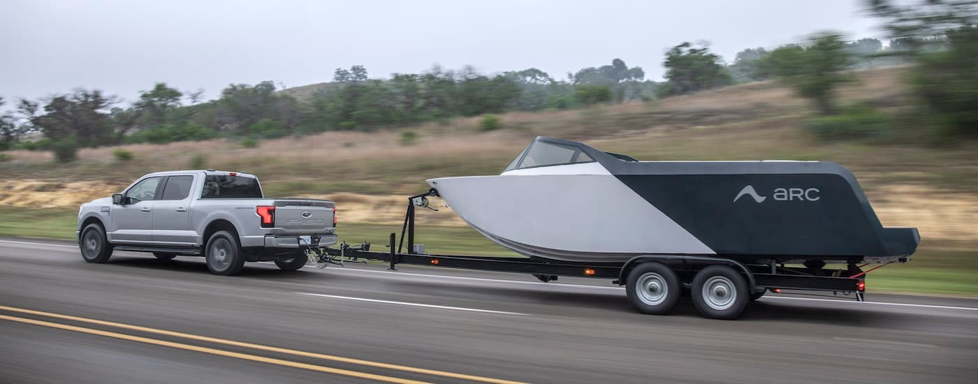 A silver 2024 Ford F-150 Lightning is shown towing a boat after leaving a dealer that sells Ford EVs.