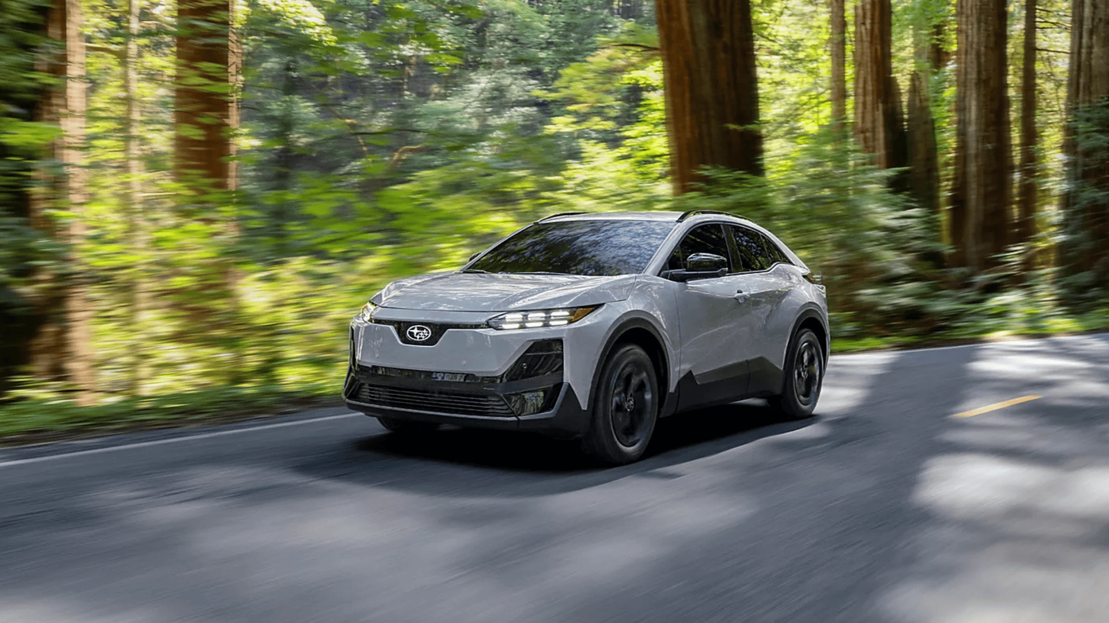 2026 Subaru Uncharted compact electric SUV in white with black wheels driving through lush green forest road in Colorado Springs CO