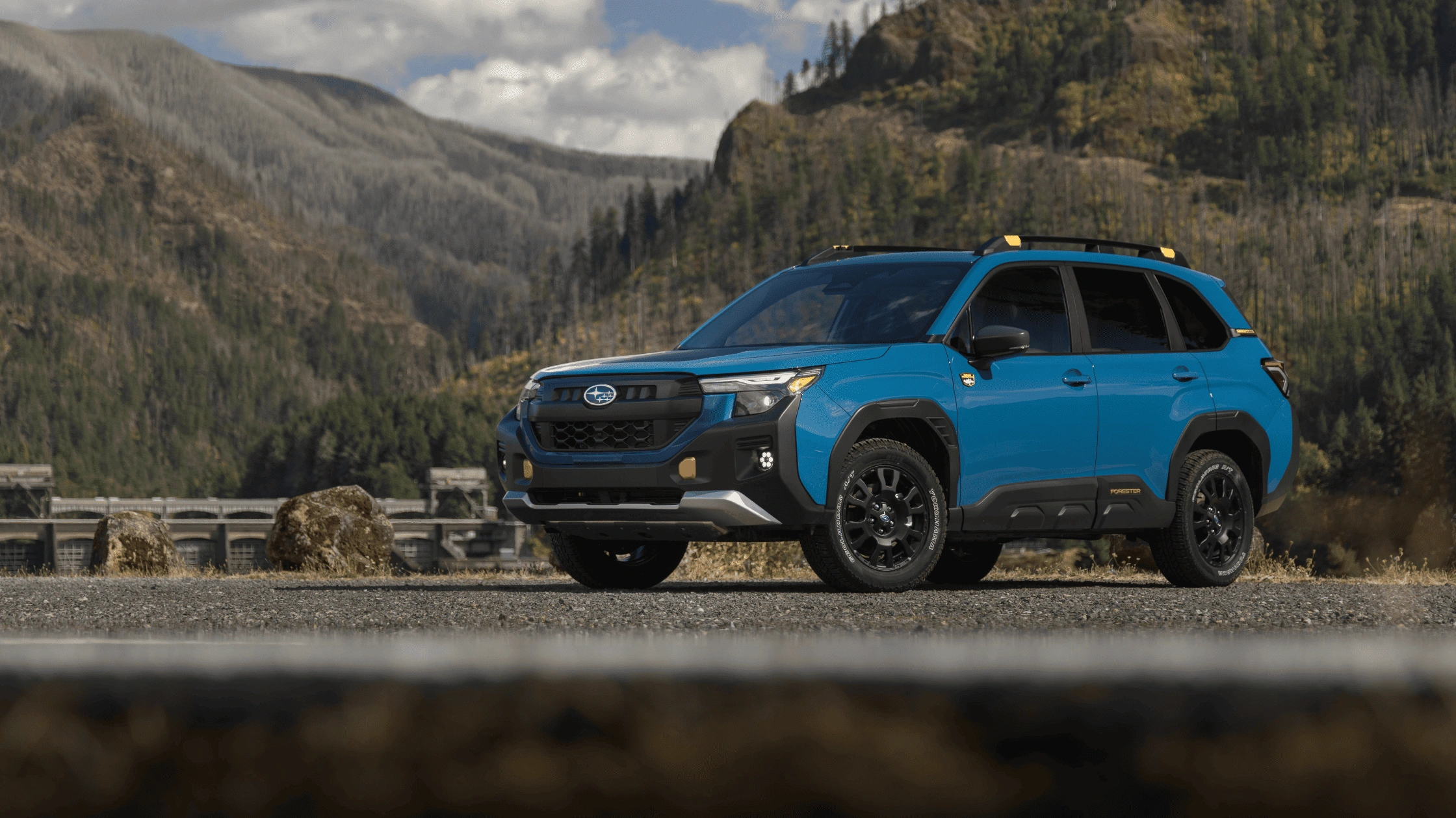 2026 Subaru Forester Wilderness in blue with yellow trim and all-terrain tires parked on rocky road with forested mountains in Pueblo CO