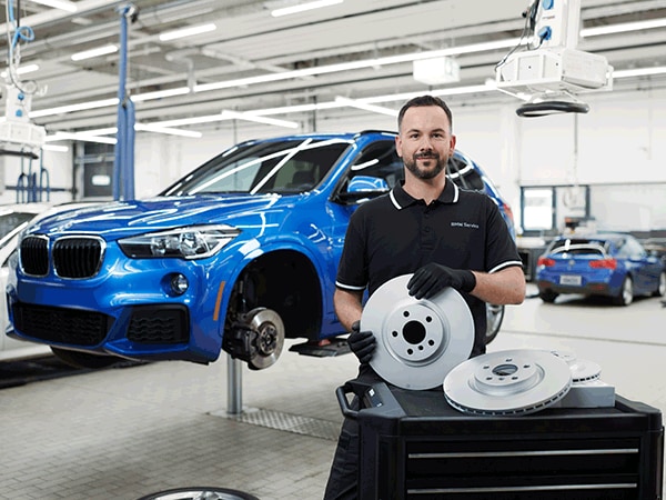 BMW technician showcasing a set of new brake rotors beside a lifted blue BMW SUV in the service department